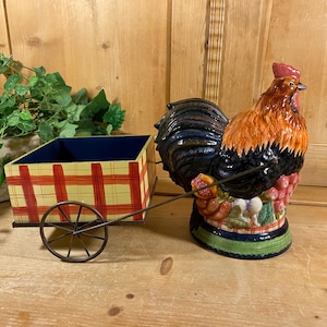 May include: A ceramic rooster figurine attached to a small, rectangular wooden cart with a yellow and red plaid pattern. The rooster features orange, black, and brown feathers, a red comb, and is positioned next to a metal wheel and handle.