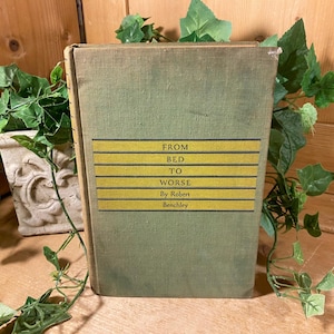 May include: A vintage book titled "From Bed to Worse" by Robert Benchley. The book has a green cover with a yellow and black title band. The book is surrounded by green ivy and a decorative pot.