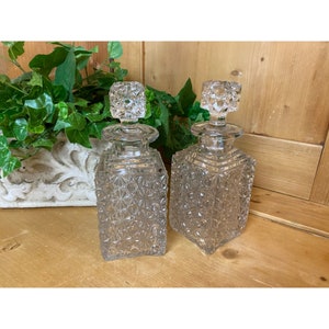 May include: Two clear glass decanter bottles with a square shape and a textured surface. The bottles have a clear glass stopper with a geometric design.