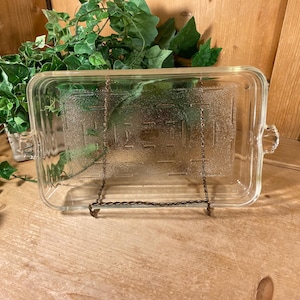 May include: A rectangular clear glass baking dish with rounded corners and integrated side handles. The base features a textured, geometric pattern of raised lines and rectangles. The dish is displayed upright on a wire stand.