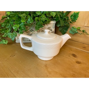 May include: White ceramic teapot with a lid and a simple design. The teapot has a rounded body and a spout on one side. The teapot is sitting on a wooden surface.