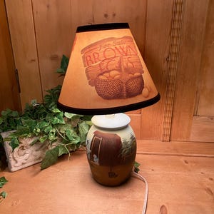 May include: A table lamp with a tan lampshade featuring a brown graphic of a basket of eggs and the text For Sale BROWN EGGS. The ceramic base is painted with a rural scene showing a small building and trees in shades of brown and green.