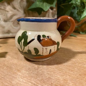 May include: A small, hand-painted ceramic pitcher with a brown handle and a cream-colored body. The pitcher features a blue rim and a painted scene of a house and trees. The handle is a warm brown color.