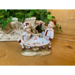 May include: A porcelain figurine of two children, a girl and a boy, holding a small white bowl with pink flowers. The children are dressed in red and white clothing and are standing on a base with a brown and green landscape.