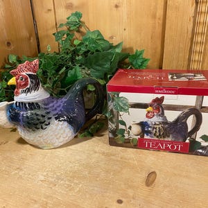 May include: A ceramic teapot shaped like a rooster, featuring a red comb, yellow beak, and blue and white plumage. The teapot is next to its original box, which also displays the rooster design. The teapot is a decorative kitchen item.