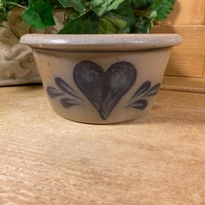 May include: A tan stoneware bowl with a wide rim and a tapered base. The exterior features a hand-painted dark blue heart design centered on the front, flanked by stylized leaf motifs on both sides of the heart, creating a symmetrical pattern.