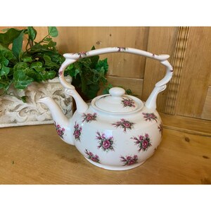May include: White ceramic teapot with a floral design featuring pink roses. The teapot has a curved body and a unique handle with a decorative design.