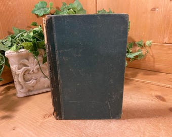 May include: A vintage, dark teal book with a worn cover and spine. The book is standing upright on a wooden surface, with a decorative white planter and greenery in the background. The book's edges show signs of age.