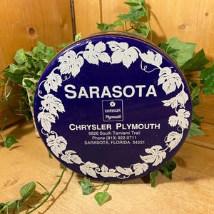 May include: A round, dark blue metal tin featuring a white grape leaf border. The text reads: SARASOTA, CHRYSLER PLYMOUTH, 6826 South Tamiami Trail, Phone (813) 922-0711, SARASOTA, FLORIDA 34231, with a Chrysler Plymouth logo in the center.
