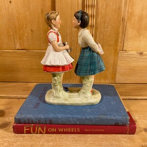 May include: A ceramic figurine of two girls facing each other, one in a white dress with a red trim and the other in a plaid skirt. The figurine is on a blue book with the words "THE OTHER SIDE" and "FUN ON WHEELS" visible.