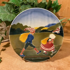 May include: A decorative ceramic plate featuring a pastoral scene with two figures walking through a landscape. The plate has a blue sky, green hills, and a small village in the background. The figures are wearing colorful clothing.