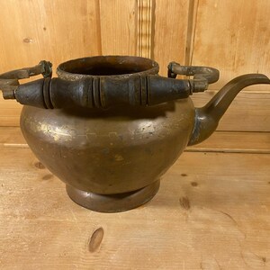 May include: Antique brass teapot with a rounded body, spout, and two wooden handles. The teapot has a dark patina and sits on a small base. The handle is made of dark wood. A vintage kitchenware item.
