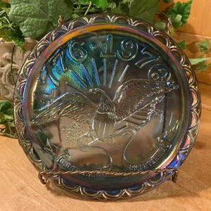 May include: Iridescent glass plate with an embossed eagle design, the year 1976, and a decorative rim. The plate is displayed on a metal stand, with a background of greenery and wood.