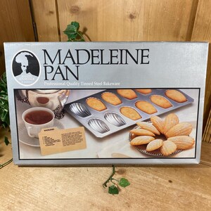 May include: A silver box with the words "MADELEINE PAN" and an illustration of a woman. The box contains a baking pan with madeleine cookies, a cup of tea, and a recipe card. The box is labeled "Professional Quality Tinned Steel Bakeware."