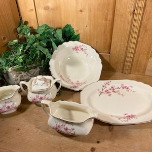 May include: A collection of vintage white porcelain serving dishes with a pink floral design. The set includes a serving platter, a bowl, a sugar bowl, and two cream pitchers. The edges are trimmed with gold. The dishes are arranged on a wooden surface.