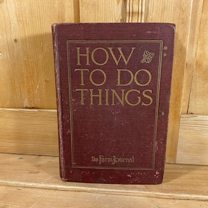 May include: A vintage book titled "HOW TO DO THINGS" with gold lettering on a maroon cover. The book has a rectangular gold border and the text "The Farm Journal" at the bottom. The book is standing upright against a wooden background.