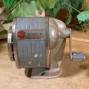 May include: A vintage Apsco Giant 51 manual pencil sharpener with a grey metal body and a silver crank handle. The front label features the text Apsco Products Inc., Apsco, Giant 51, Beverly Hills, Calif., Rockford, Ill. U.S.A., Type III Gear Assembly.