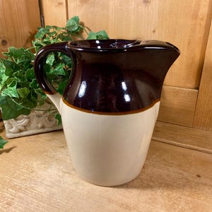 May include: A ceramic pitcher featuring a two-tone design with a glossy dark brown upper section and a smooth cream-colored lower body. The vessel includes a curved handle on the side and a wide, integrated pouring spout at the top rim.