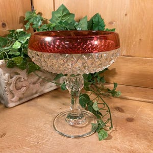 May include: A vintage champagne glass with a ruby red rim and a clear, diamond-cut bowl. The glass sits on a clear stem and base. The glass is likely made of crystal and is a decorative piece.