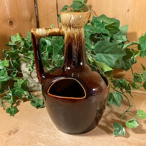 May include: A vintage brown ceramic pitcher with a handle and a unique cut-out design. The pitcher has a glossy finish and a lighter-colored rim. The background includes green foliage and a wooden surface.