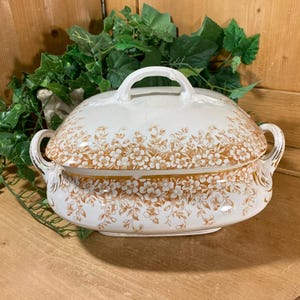 May include: White porcelain serving dish with a lid and brown floral pattern. The dish has two handles on the sides.