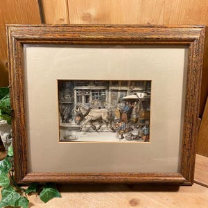 May include: Framed artwork depicting a horse-drawn carriage on a cobblestone street. The artwork is in a brown wooden frame with a beige mat. The scene includes figures and buildings, creating a detailed historical illustration.