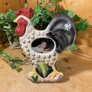 May include: A ceramic rooster-shaped picture frame featuring a white body with black feather patterns, a bright red comb and wattle, and a dark gray tail. The center of the rooster contains an oval opening displaying a photograph of a baby.