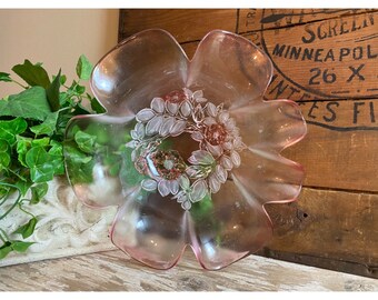 May include: A pink glass flower-shaped dish with a floral design in the center. The dish has six petals and a detailed wreath of leaves and flowers in the center. The background includes a wooden crate and greenery.