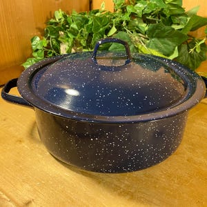 May include: A navy blue enamelware pot with a lid. The pot has a speckled white pattern and two side handles. The lid has a handle on top. The pot is sitting on a wooden surface.