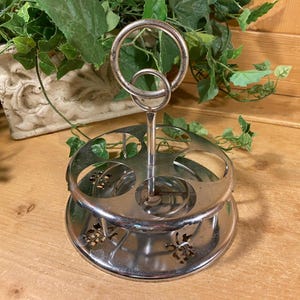 May include: A silver-colored egg holder with a circular handle and six egg cup holders. The base has a decorative floral design. The holder is made of metal and sits on a wooden surface.