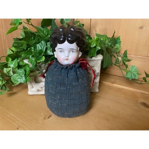 May include: A vintage porcelain doll head with black hair and painted blue eyes, wearing a dark blue fabric bag with a red ribbon.