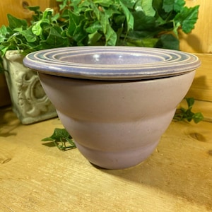 May include: A lavender-colored ceramic bowl with a matching lid. The bowl has a rounded shape with a series of concentric rings on the lid. The rim of the lid is a lighter shade of lavender. The bowl is sitting on a wooden surface.