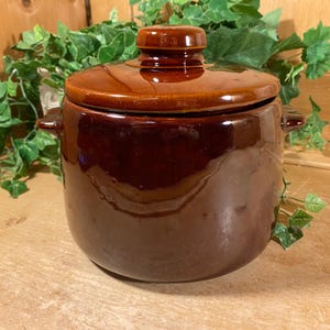 May include: A brown ceramic bean pot with a lid. The pot has a glossy finish and two small handles. The lid has a knob on top. The pot is sitting on a wooden surface.