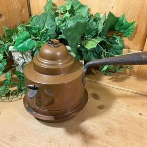 May include: A copper tea kettle with a long, dark brown handle. The kettle has a round body and a lid with a decorative knob. The kettle is sitting on a wooden surface.