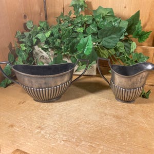 May include: Two antique silver-toned serving dishes. The larger dish has two handles and a wide opening. The smaller dish has a spout and a single handle. Both have vertical ribbed detailing.