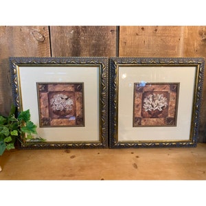May include: Two framed floral prints with white flowers in a vase on a brown background. The frames are ornate and have a gold finish.