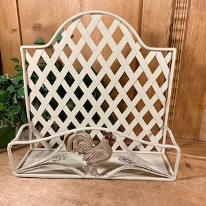 May include: A cream-colored metal wall-mounted rack featuring a lattice-patterned back panel and a front storage shelf. The center of the shelf is decorated with a small, three-dimensional figurine of a rooster in a muted brown and tan finish.
