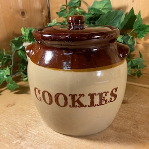 May include: A vintage ceramic cookie jar with a brown lid and base, and a tan body. The word "COOKIES" is printed in brown on the front. The jar has two small handles and a knob on the lid. The background includes green foliage.