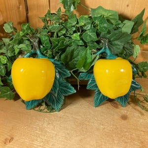 May include: Two ceramic apple-shaped decorations with bright yellow bodies and green stems and leaves. The apples are set against a backdrop of green foliage, creating a vibrant and naturalistic display. The items are likely decorative accents.