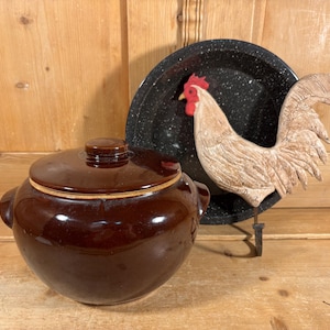 May include: A dark brown glazed ceramic bean pot with a matching lid and two side handles, positioned next to a black speckled enamelware plate featuring a three-dimensional ceramic rooster figure with a red comb and white feathers.