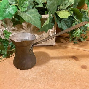 May include: A vintage copper coffee pot with a long handle. The pot is made of metal and has a dark brown patina. It is sitting on a wooden surface.
