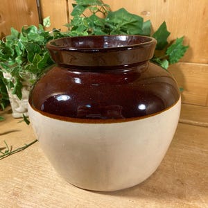May include: A round ceramic crock featuring a two-tone design with a glossy dark brown glazed upper section and a matte cream-colored lower body. The vessel has a wide, open mouth with a thick, slightly flared rim and a smooth, rounded silhouette.