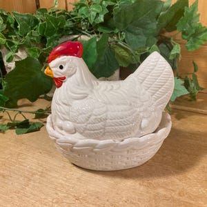 May include: A white ceramic hen-shaped container with a red comb and wattle, and an orange beak. The hen sits atop a woven basket-like base. Green foliage is in the background, and the piece is on a wooden surface.