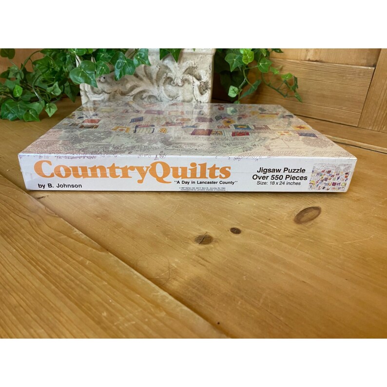 Może przedstawiać: Białe pudełko z puzzlem Country Quilts, "A Day in Lancaster County" autorstwa B. Johnsona. Pudełko jest białe z pomarańczowym tekstem i obrazkiem kołdry na przodzie. Puzzle zawiera ponad 550 element&oacute;w i ma wymiary 46 x 61 cm.