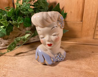 Peut inclure: Un vase en céramique en forme de tête représentant un visage stylisé aux yeux fermés, joues roses et lèvres rouges. La figurine a une coiffure claire avec une fleur bleue et porte un vêtement bleu avec des bordures dorées et une petite fleur dorée.