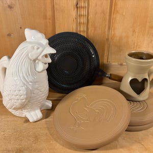 May include: A collection of kitchenware items, including a white ceramic rooster pitcher, a black perforated pan, a tan ceramic oil warmer with a heart-shaped cutout, and two tan ceramic trivets with rooster designs.