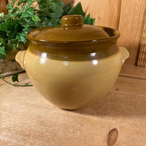 May include: A round ceramic pot with a glossy finish, featuring a light tan base and a darker brown glazed upper section and lid. The pot includes two small side handles and a central knob on the lid, showcasing a two-tone color scheme.