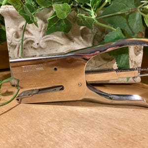 May include: Vintage chrome and wood stapler. The stapler has a silver handle and a wooden base. The brand name "LEITZ" is visible on the side. The stapler is on a wooden surface with greenery in the background.