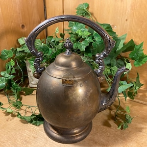 May include: Antique brass teapot with a silver-colored handle and spout. The teapot has a round body and a hinged lid. The handle is arched and ornate. The spout is curved. The teapot sits on a round base. Decorative kitchenware.