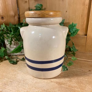 May include: A cream-colored ceramic jar with a wooden lid. The jar has two dark blue stripes and two small handles. The jar is sitting on a wooden surface with green foliage in the background.